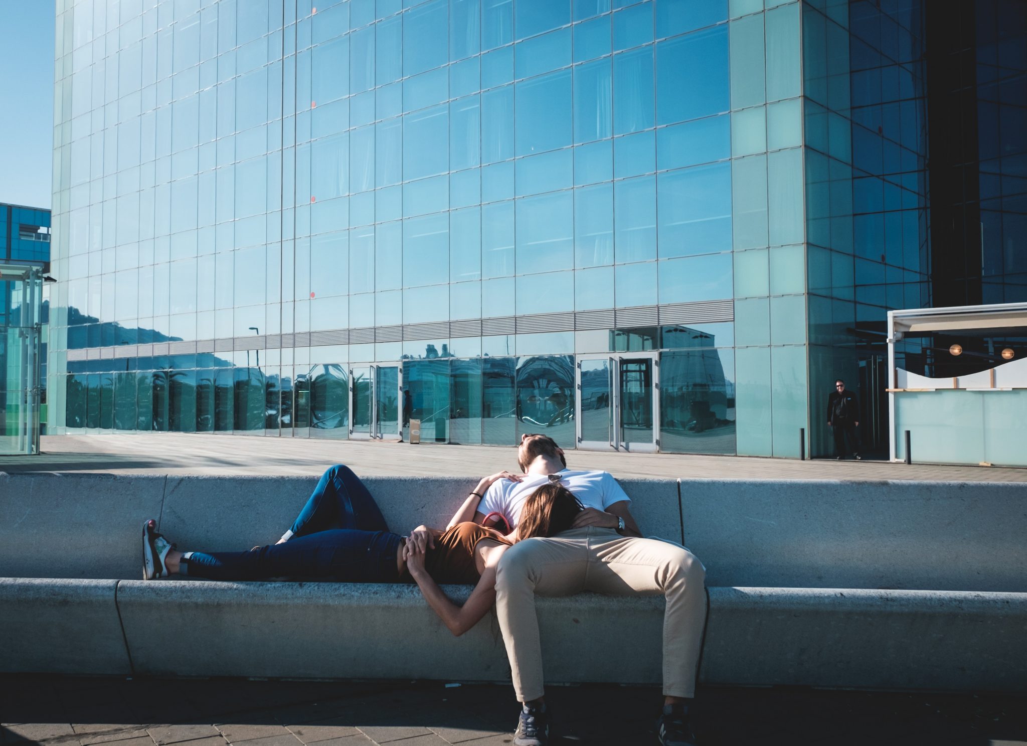 couple sleeping in new york sitting on a long stone bench with the woman resting her head in man's lap with glossy skyscraper background and blue sky
