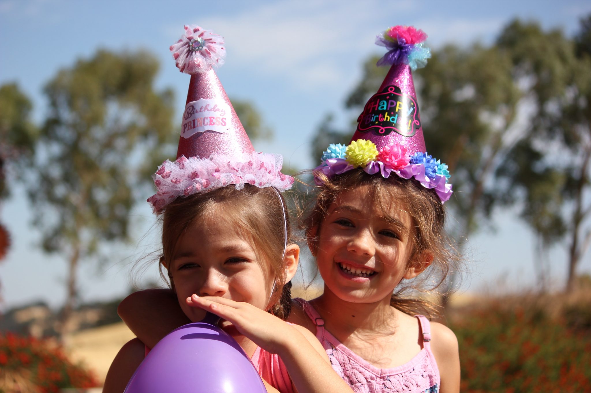 two girls in party hats one blowing up a purple balloon, outside in the sun