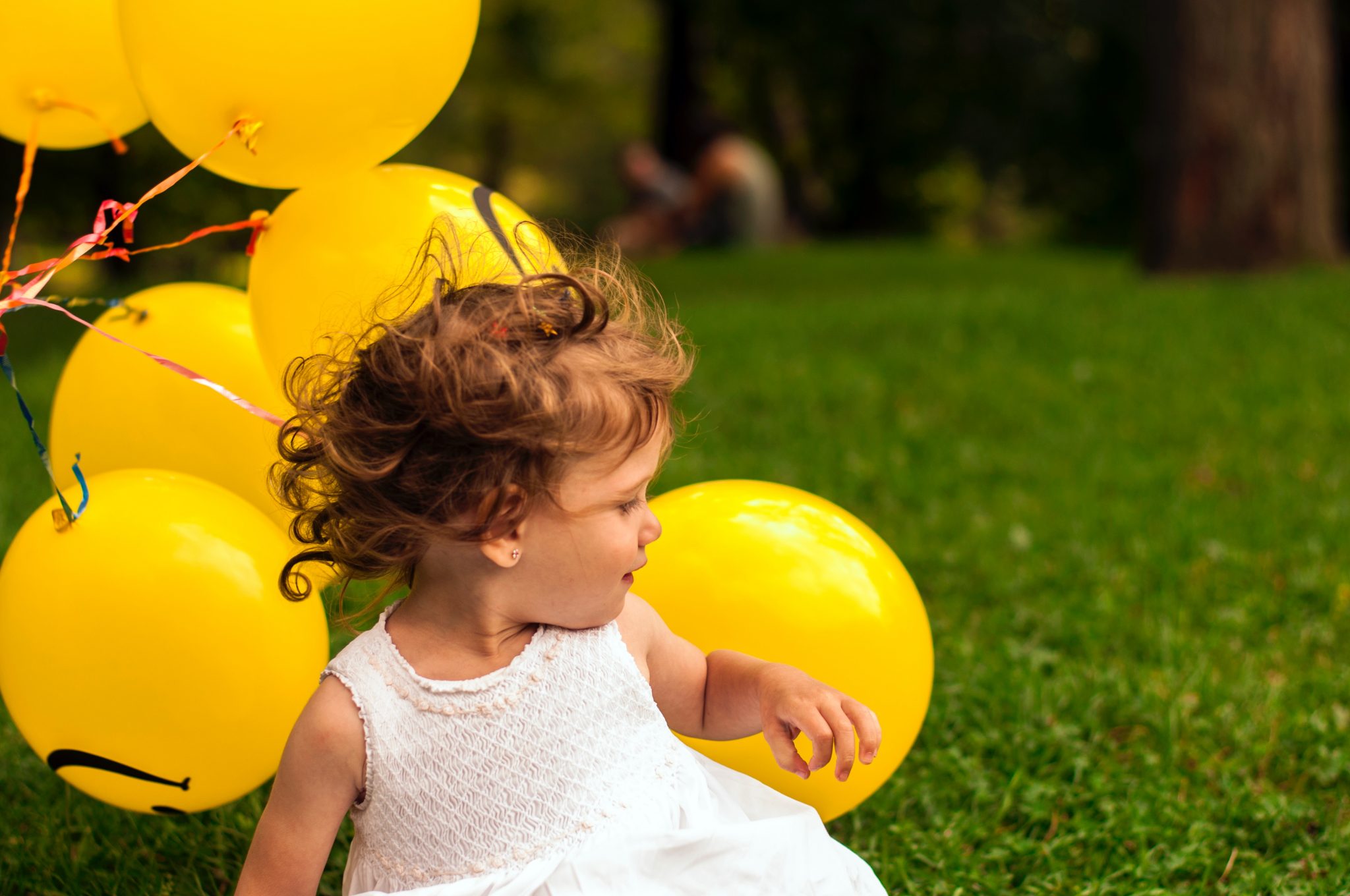Little girl in white dress on grass background next to bunch of yellow balloons